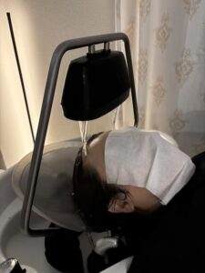 Person lying with head under a curved salon shampoo faucet, water flowing over their hair, a cloth covering their eyes.