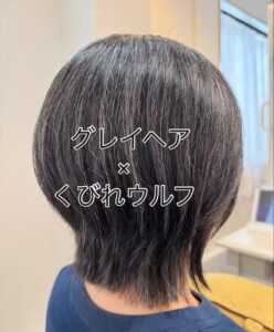 Back view of a person with short, silver-gray hair in a tapered wolf cut, in a salon setting. Overlay text reads 'グレイヘア × くびれウルフ'