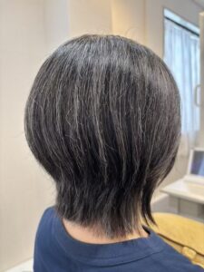 Back view of a person with short layered gray hair, wearing a blue shirt, in a bright indoor space (likely a salon or clinic).