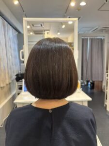 Back view of a person with a sleek dark brown bob in a salon chair, mirrors and lighting in the background, showcasing a polished cut.